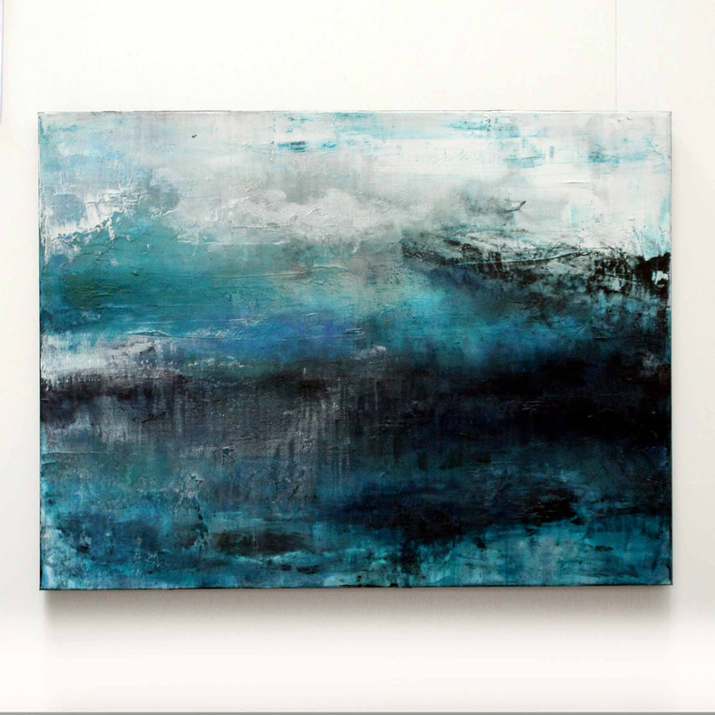 An abstract canvas painting featuring textured brushstrokes of teal, blue, black, and white, with a hazy, atmospheric composition displayed on a plain white wall in the artists studio.