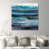 Abstract seascape canvas wall art painting featuring horizontal brushstrokes in shades of blue, teal, navy, and grey, displayed above a white sofa with neutral throw pillows in a modern living room setting.