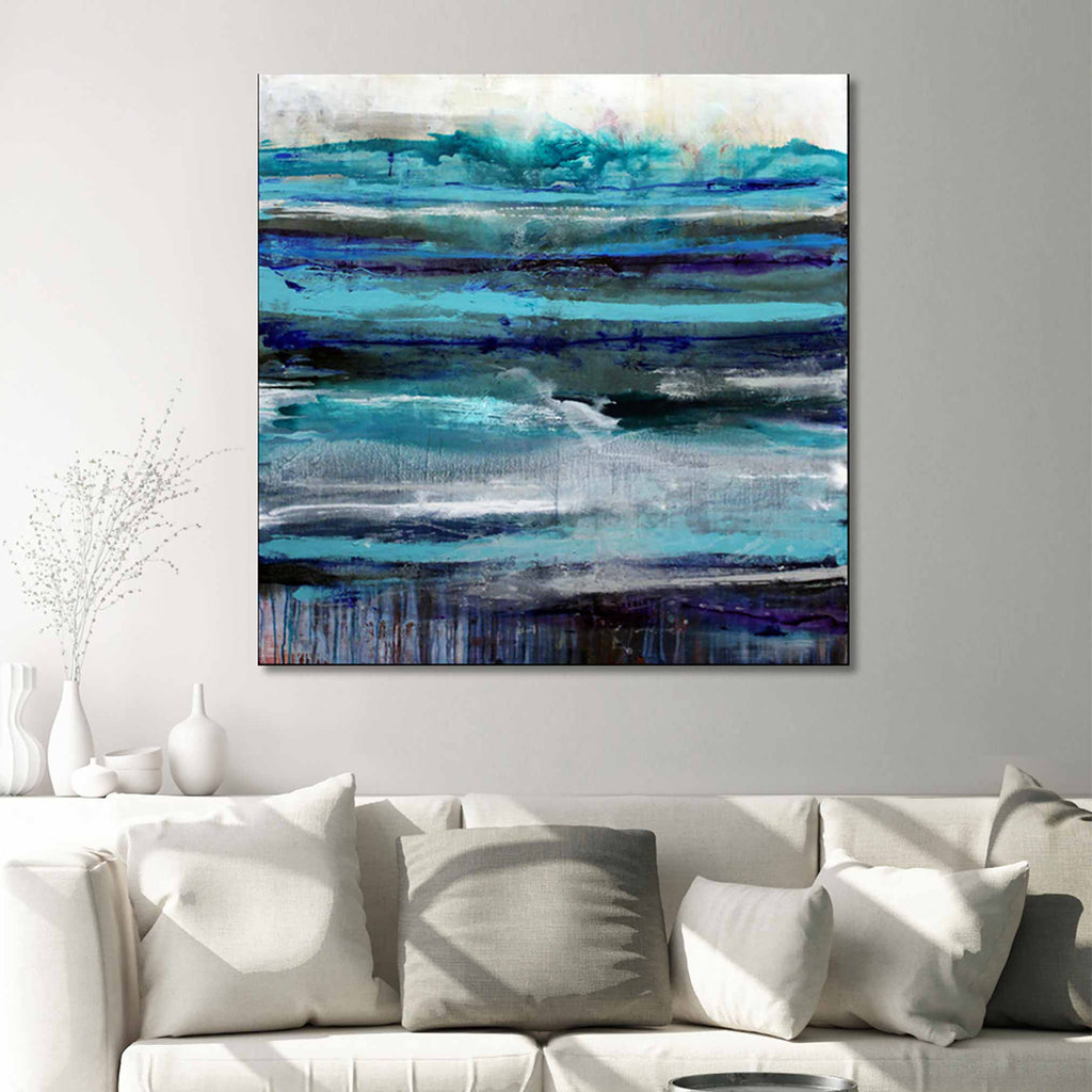 Abstract seascape canvas wall art painting featuring horizontal brushstrokes in shades of blue, teal, navy, and grey, displayed above a white sofa with neutral throw pillows in a modern living room setting.