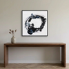 Orbital - A square framed abstract wall art painting hangs above a wooden console table against a light beige wall. The artwork features a black, gray, and white circular brushstroke design.