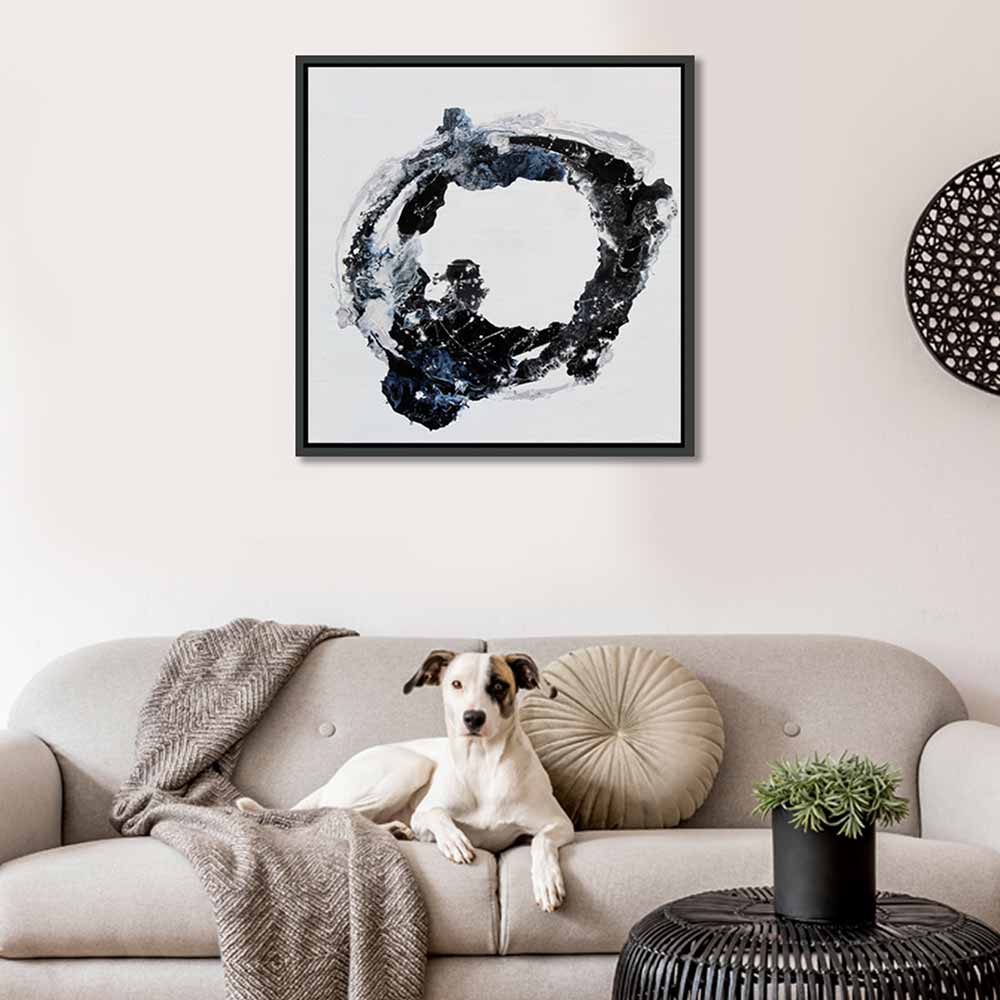 A framed abstract wall art painting orbital with a black and blue brushstroke design on a white background, hanging above a beige sofa in a living room. A white dog lies on the sofa, with a throw blanket, round cushion, and small potted plant nearby.