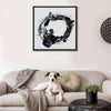 A framed abstract wall art painting orbital with a black and blue brushstroke design on a white background, hanging above a beige sofa in a living room. A white dog lies on the sofa, with a throw blanket, round cushion, and small potted plant nearby.