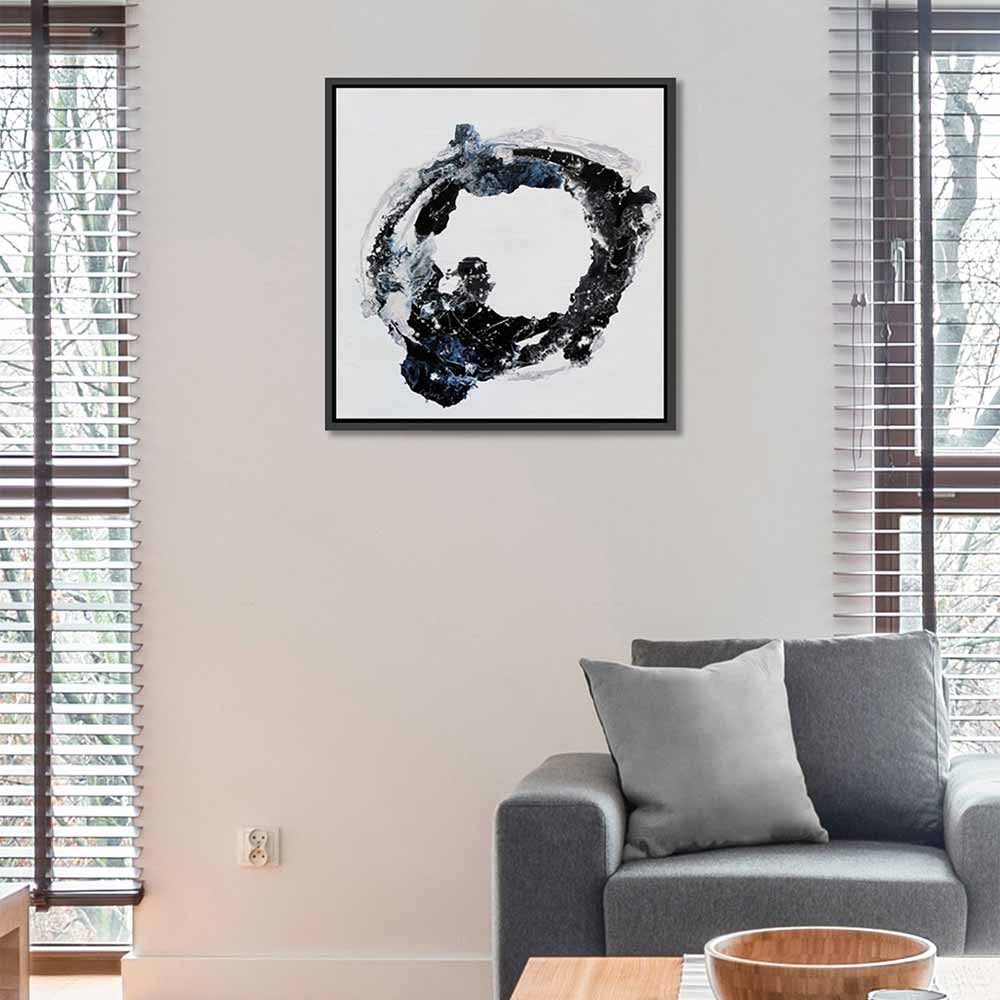 A framed abstract black, white, and blue circular painting called Orbital hanging on a beige wall above a grey sofa in a modern living room setting.