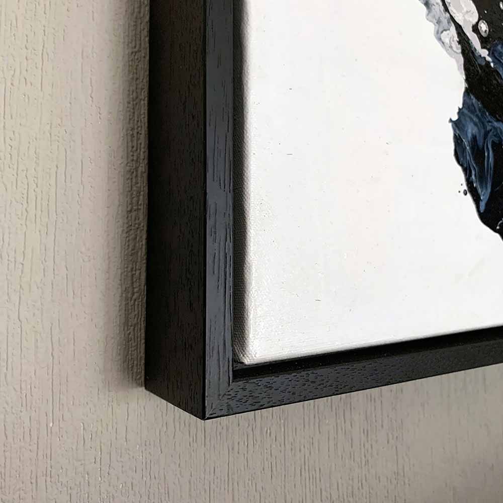 Close-up of a framed wall art painting Orbital showing a dark wood frame with a white mat and part of a black-and-white abstract painting on a textured beige wall.