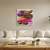 A colorful abstract canvas painting with vibrant shades of purple, pink, orange, yellow, and green, displayed in a white frame on a beige wall above a modern white sofa in a living room setting.