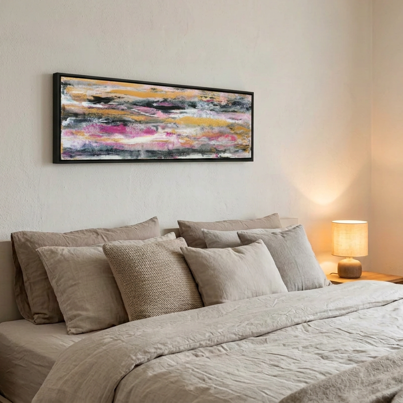 A modern bedroom scene featuring a wide rectangular abstract canvas painting with black, gray, yellow, and pink brushstrokes, hung on a textured off-white wall above a bed with neutral beige and gray linens and pillows, with a lit bedside lamp casting a warm glow.