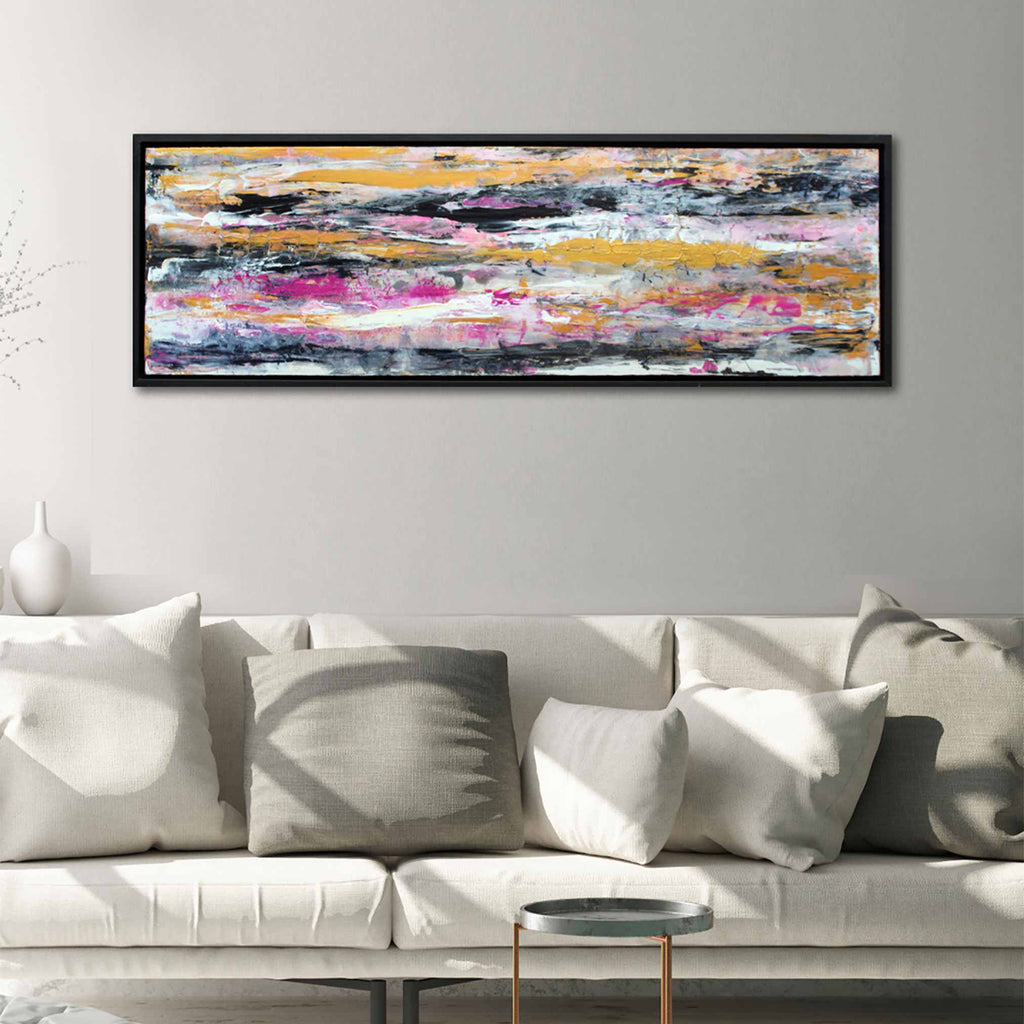 A black framed modern contemporary wave painting by British artist Andy Watt horizontal waves in gold pink, black, white and grey, hanging on a light grey wall with cream couch and cushions underneath.