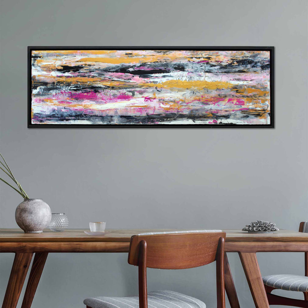 A black framed abstract canvas wave painting of blending horizontal brush strokes in gold pink, black, white and grey, hanging on  a light grey wall with dinning table and chairs underneath.