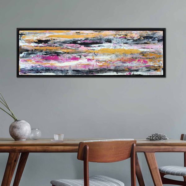 A black framed abstract canvas wave painting of blending horizontal brush strokes in gold pink, black, white and grey, hanging on  a light grey wall with dinning table and chairs underneath.