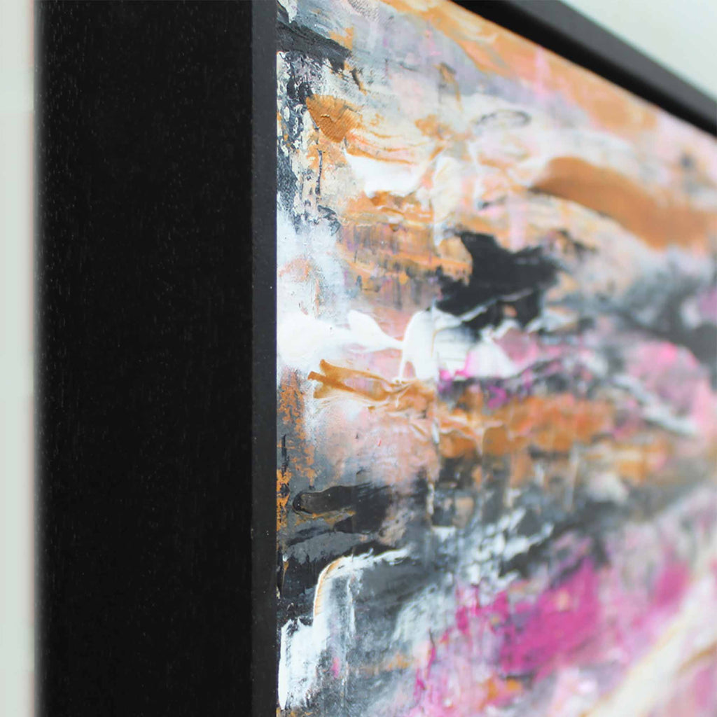 Close-up of a framed abstract painting showing a thick black frame and textured brushstrokes in white, gray, black, gold, and pink.