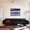 An abstract canvas painting with horizontal bands of blue, pink, red, and black hangs on a white wall above a black leather sofa in a modern living room setting.