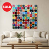 Vintage spheres No 10 - A square abstract canvas painting featuring a sold circle in red with white text top left of image, A colourful grid of painted blocks and circles in various hues, displayed on a beige wall above a modern white sofa in a bright, minimalist living room.