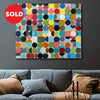 A colourful abstract canvas painting featuring a grid of multi-coloured circles on a light background, shown hanging on a dark grey wall above a grey sofa with brown and blue pillows. A red circular 'SOLD' badge is overlaid in the top-left corner.