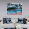 An abstract canvas wall art painting featuring vibrant blue, pink, black, and white brushstrokes, displayed above a modern white sofa with decorative blue and black pillows in a living room setting.