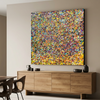 Summer Showers  is a large square abstract canvas painting featuring a dense, colourful confetti-like pattern in yellow, blue, red, green, and orange, displayed on a beige wall above a wooden credenza in a modern dining room setting.
