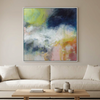 Summer Showers - A large framed abstract canvas painting with a colourful mix of blue, green, yellow, pink, and white paint strokes, hanging on a beige wall above a modern white sofa with a wooden coffee table in front.