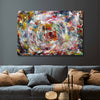 A large rectangular abstract canvas painting with a swirling multi colour paint pattern hangs on a dark grey wall above a blue-grey sofa with brown pillows in a living room setting.