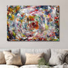 A square abstract canvas wall art painting featuring a vibrant, swirling paint pattern in multiple colours, displayed on a light grey wall above a modern sofa with neutral coloured cushions.