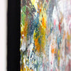 Close-up of a framed abstract painting with a thick black frame and a multicolored paint-splattered design featuring yellow, orange, green, blue, white, and red tones.