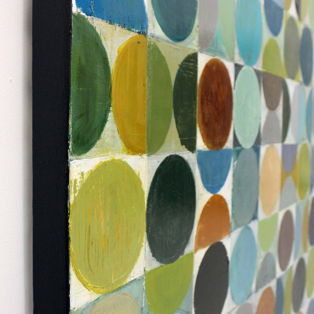 Close-up of a framed abstract geometric painting featuring painted circles in green, yellow, blue, brown, gray, and beige on a white background, with a black frame visible against a white wall.