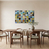 Vintage Spheres No 2 - A large rectangular wall art painting with an abstract pattern of overlapping circles and arcs in blue, teal, olive, mustard, brown, black, and grey, displayed above a wooden dining table with chairs in a bright neutral dining room.