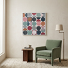 A large square framed wall art painting with an abstract pattern of colourful circles and semicircles hangs above a green armchair in a styled living room setting.