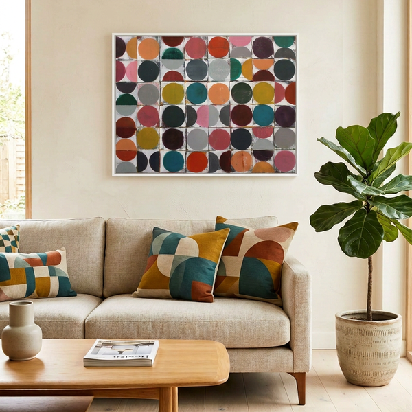 A framed abstract art painting featuring a colourful grid of circles hanging on a beige wall above a light beige sofa with patterned throw pillows in a bright, modern living room.