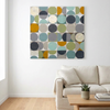 A square canvas wall art painting featuring an abstract geometric pattern of overlapping circles in muted teal, olive green, mustard yellow, navy, grey, and cream, displayed on a white wall above a modern beige sofa in a bright living room.