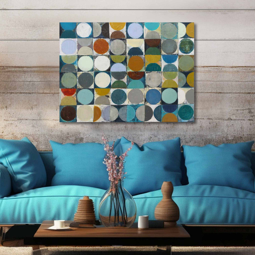 A square abstract canvas print with a grid of colorful circles in blue, teal, yellow, brown, and gray, hanging on a rustic wood-paneled wall above a turquoise sofa with decorative vases and pillows.