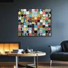 A square abstract canvas print featuring a colorful grid of overlapping circles in many hues, displayed on a dark gray wall in a modern living room setting with a fireplace, coffee table, and black leather chair.