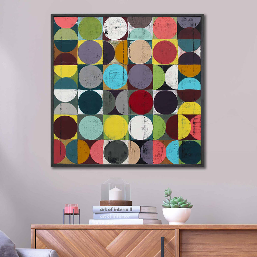 A square black framed abstract artwork featuring a grid of colourful circles and half-circles in many bright tones on a grid background the painting is hanging on a light grey wall.