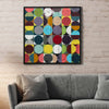 A square framed abstract artwork featuring a grid of colourful circles and half-circles in many vibrant and bright tones on a brick wall with grey couch underneath.