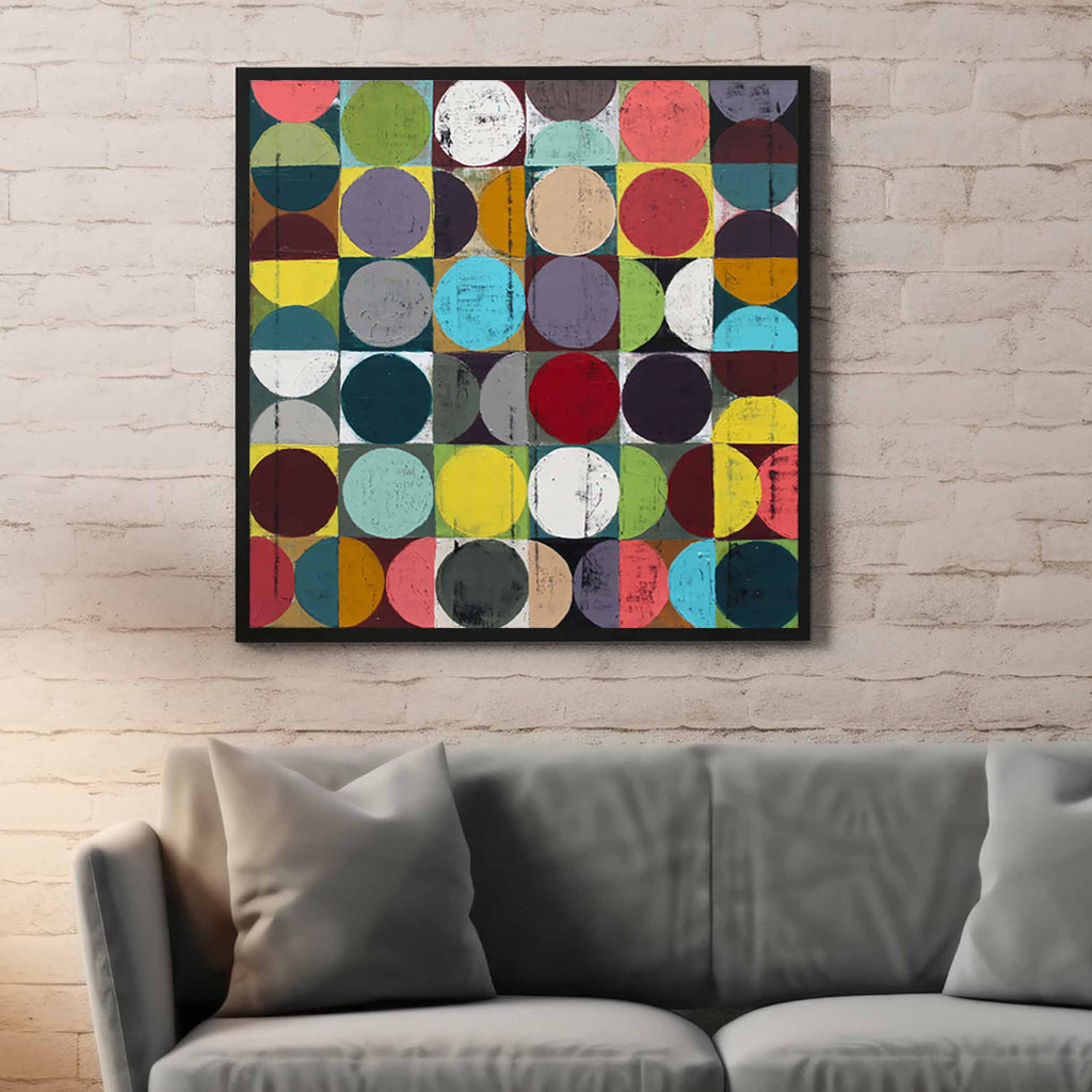 A square framed abstract artwork featuring a grid of colourful circles and half-circles in many vibrant and bright tones on a brick wall with grey couch underneath.