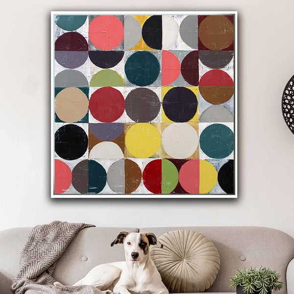 A square framed wall art print featuring a colorful geometric pattern of overlapping circles in red, yellow, teal, black, beige, and brown, displayed above a beige sofa with a white dog and a small plant.