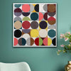 A square framed abstract wall art print featuring a grid of colorful circles and half-circles in red, yellow, blue, green, pink, black, and beige, hung on a teal wall above a white chair with pink flowers in a vase nearby.