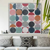 A framed square wall art print with a grid of painted circles in muted pink, teal, navy, gray, orange, and red tones, displayed above a gray sofa with patterned pillows in a styled living room.