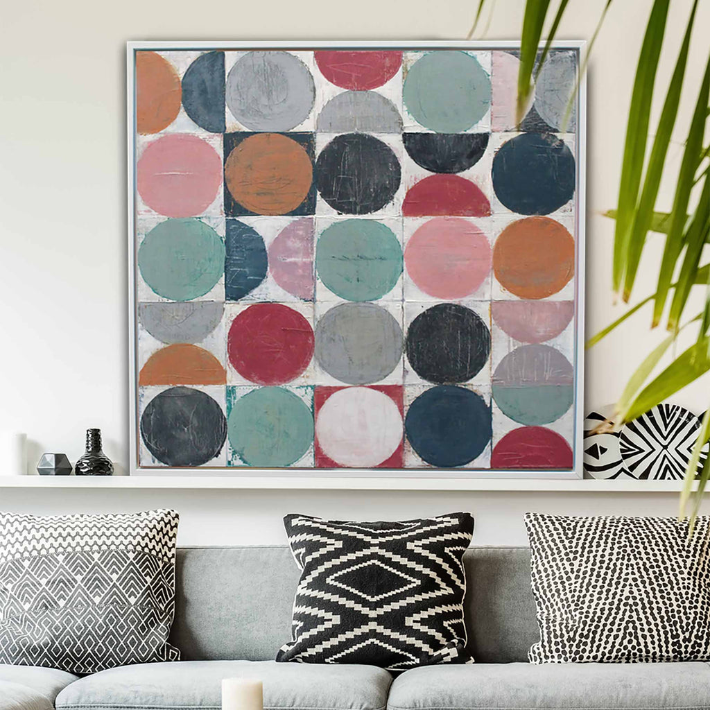 A framed square wall art print with a grid of painted circles in muted pink, teal, navy, gray, orange, and red tones, displayed above a gray sofa with patterned pillows in a styled living room.