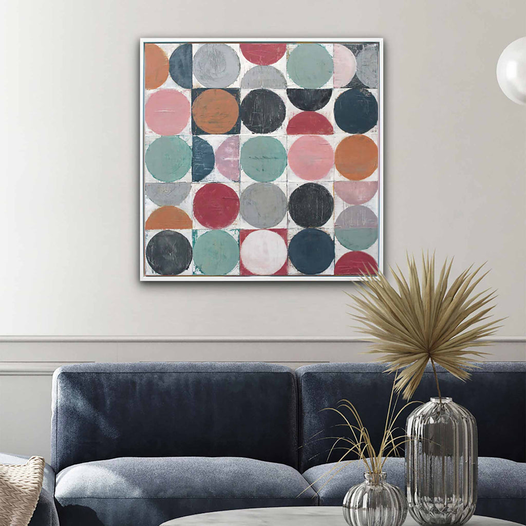 A square framed wall art print featuring a grid of colorful circles in muted pink, teal, navy, orange, gray, and red tones, displayed above a dark blue sofa in a styled living room.