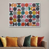A framed abstract art print featuring a grid of colorful circles in earthy tones, displayed on a beige wall above a sofa with mustard, green, and rust-colored throw pillows.