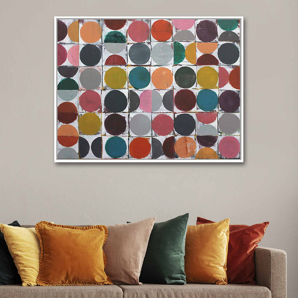 A framed abstract art print featuring a grid of colorful circles in earthy tones, displayed on a beige wall above a sofa with mustard, green, and rust-colored throw pillows.