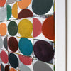 Close-up of a colorful abstract wall art piece showing painted circles and arcs in orange, yellow, red, teal, purple, green, and gray on a white grid, with a white frame edge visible on the right.