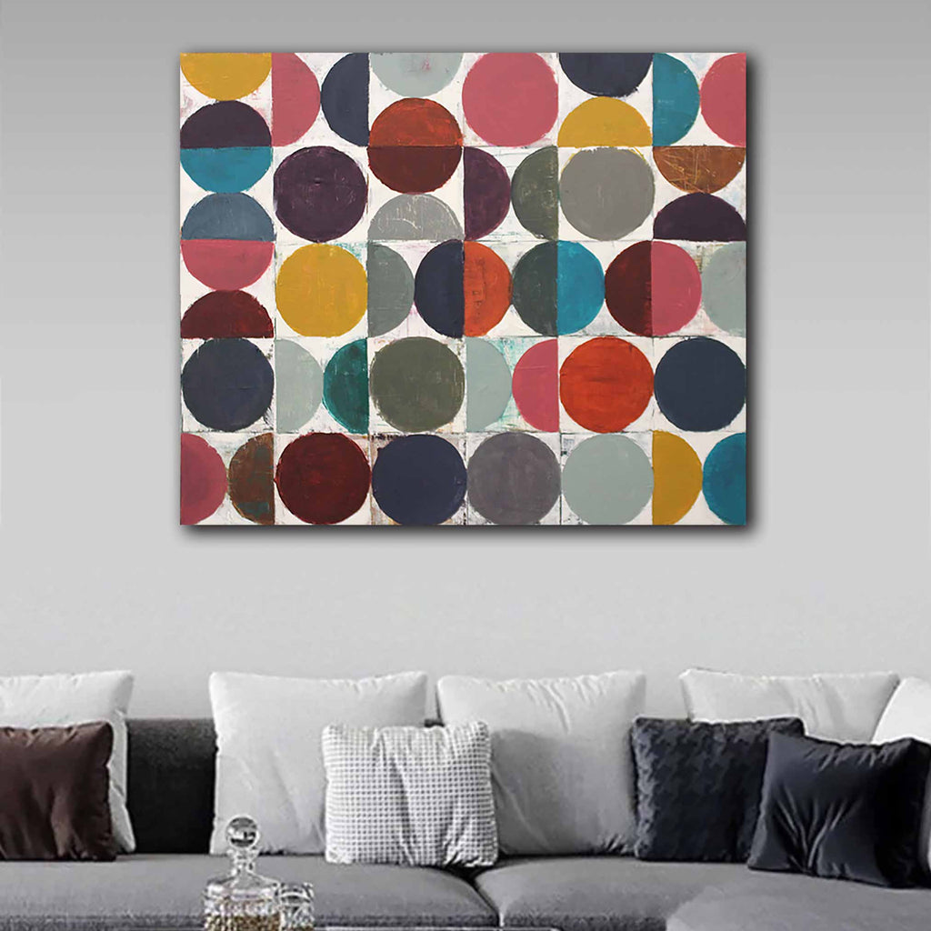 A square canvas wall art painting featuring a colourful geometric pattern of overlapping circles in muted red, mustard yellow, teal, navy, olive, and grey on a white background, displayed above a grey sofa with white and dark accent pillows.