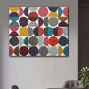 A square canvas wall art painting featuring a modern geometric pattern of overlapping circles and half-circles in multiple colours on a white background, hung on a light grey wall in a styled interior setting.