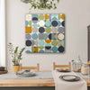 A square canvas wall art painting featuring a grid of colourful circles in teal, mustard yellow, olive green, navy, grey, and cream, hanging on a light grey wall above a wooden dining table with plates and a runner.