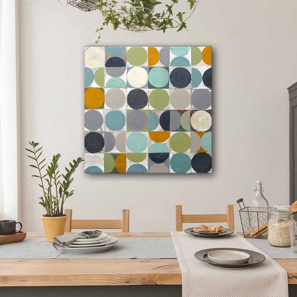 A square canvas wall art painting featuring a grid of colourful circles in teal, mustard yellow, olive green, navy, grey, and cream, hanging on a light grey wall above a wooden dining table with plates and a runner.