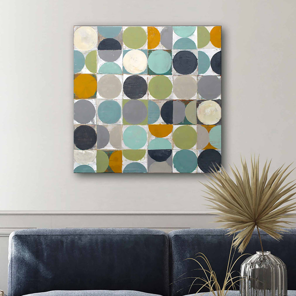 A square abstract canvas hand pain featuring a grid of colourful circles in teal, olive green, mustard yellow, navy, grey, and cream, displayed on a light grey wall above a dark blue sofa with a glass vase and dried palm leaf in the foreground.ed