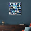 A square framed abstract wall art painting featuring a geometric pattern of overlapping circles in shades of blue, teal, grey, black, and white, displayed on a dark blue wall above a wooden console table.