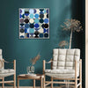 A square framed wall art painting with an abstract geometric pattern of overlapping circles in shades of blue, navy, black, grey, and cream, displayed on a dark teal wall in a modern living room setting with two light wood armchairs and a small side table.