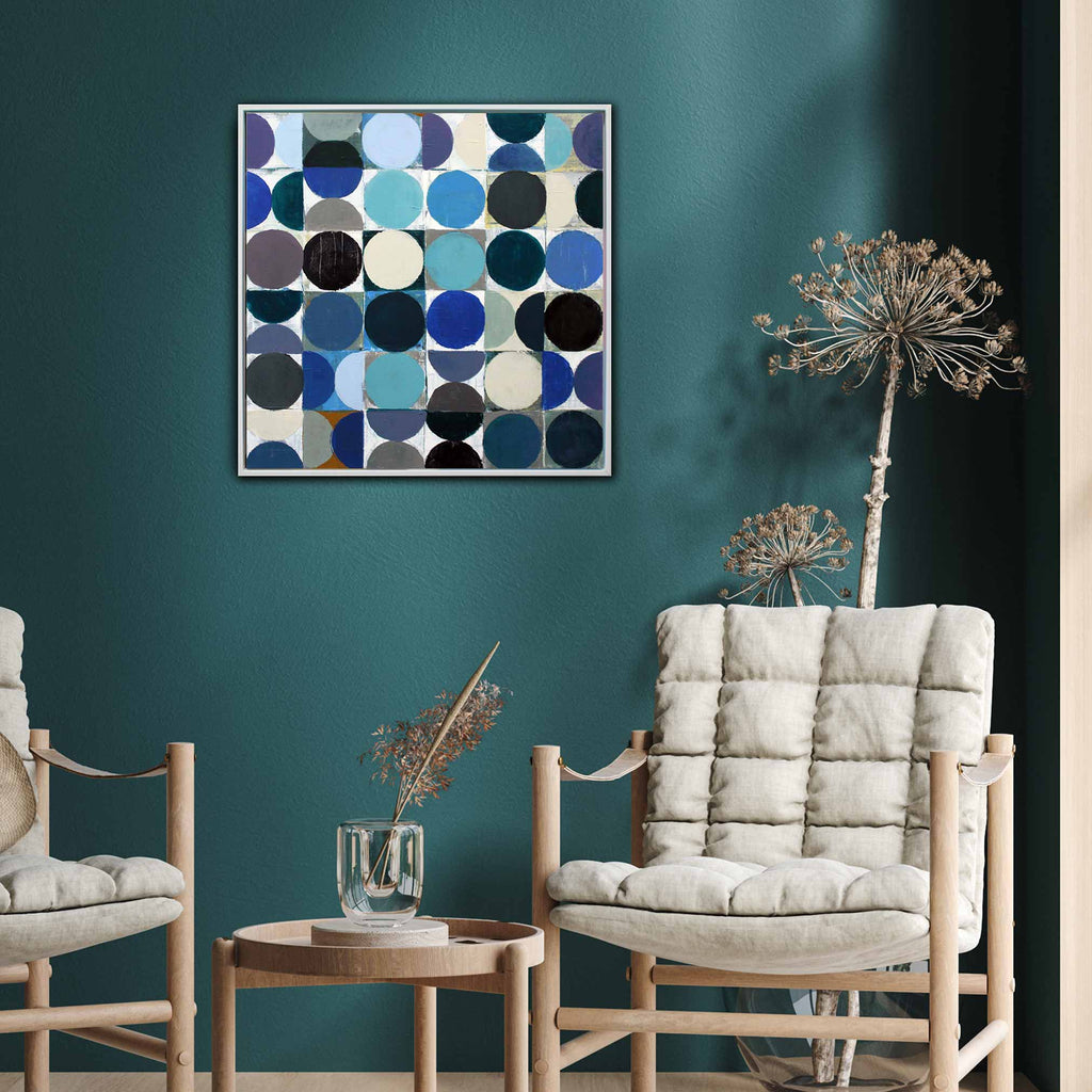 A square framed wall art painting with an abstract geometric pattern of overlapping circles in shades of blue, navy, black, grey, and cream, displayed on a dark teal wall in a modern living room setting with two light wood armchairs and a small side table.